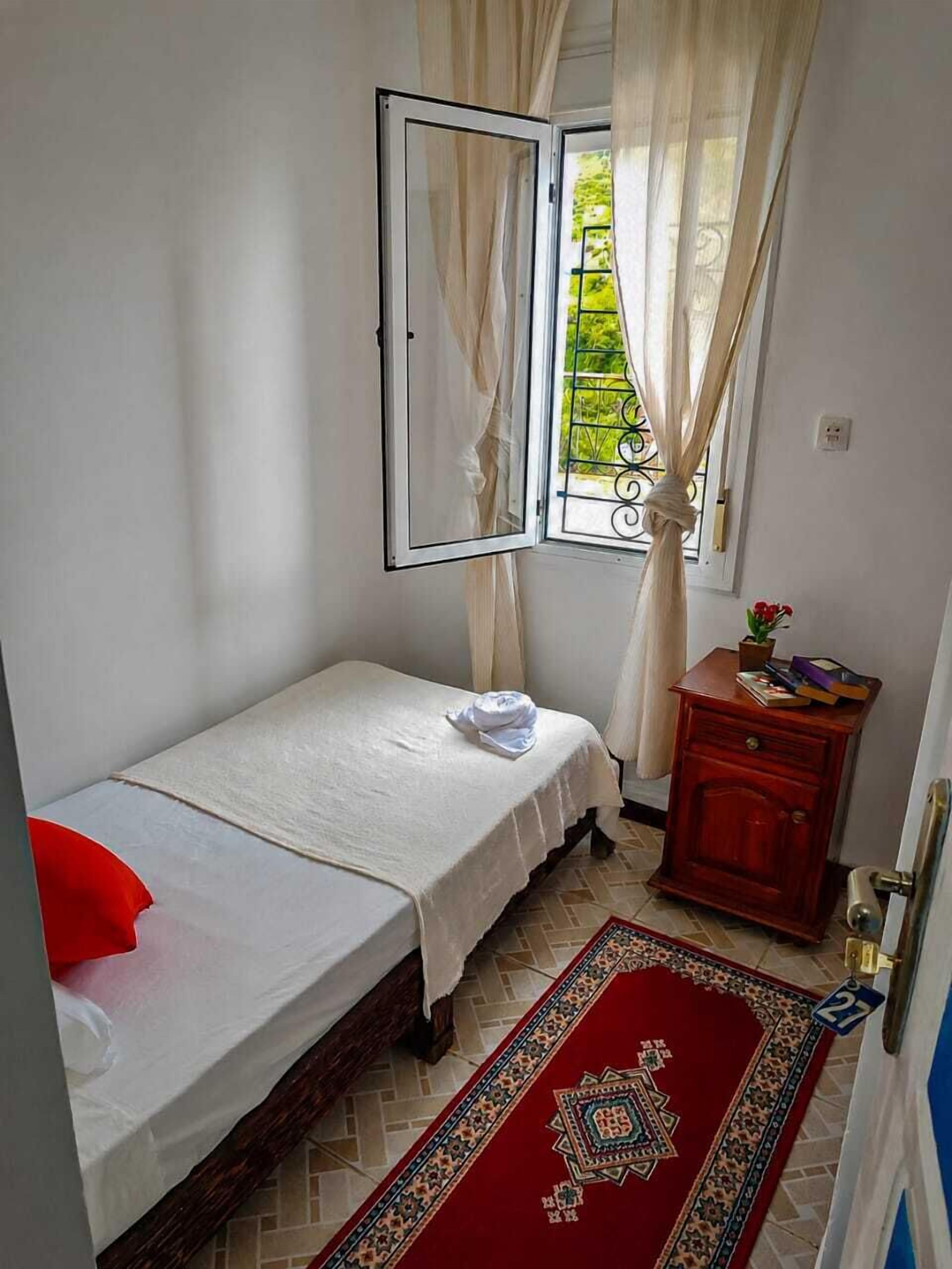 A small bedroom with a single bed, a window with curtains, a wooden nightstand, and a patterned rug.