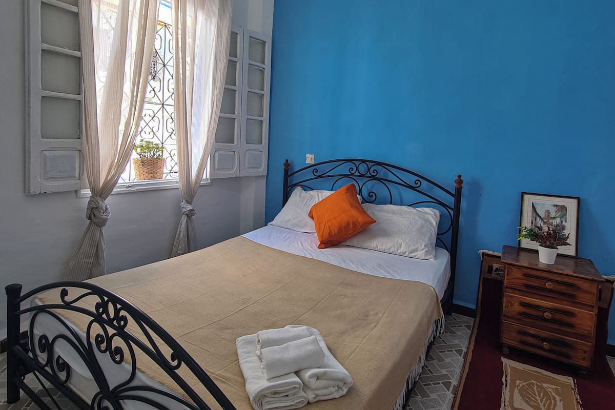 Un dormitorio con cama, mesita de noche, lámpara, ventana con cortinas y pared azul.