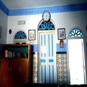 A room with a wooden bookshelf, a clock on the wall, a door with blue and white stripes, and a window with a blue pattern. - Hostal La Joya (Chefchaouen)