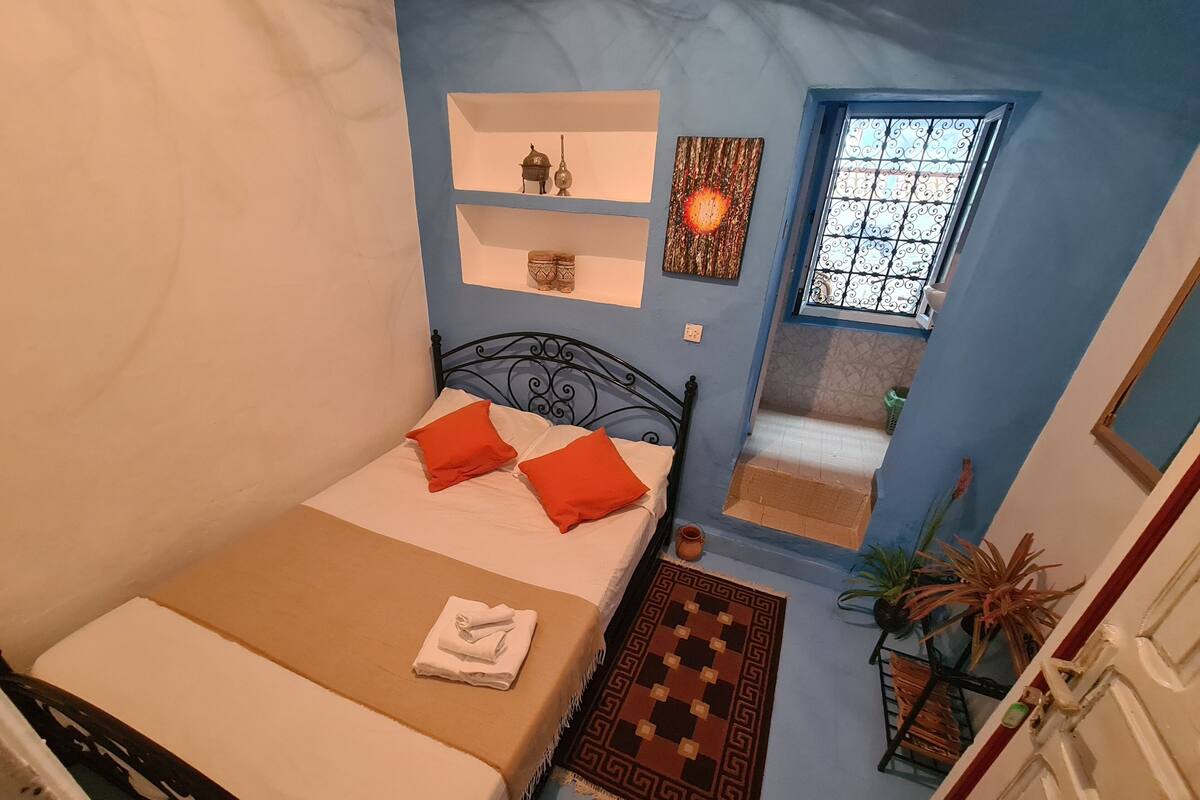 A bedroom with a bed, pillows, a small shelf with decorative items, a window, and a door.