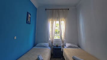 A room with two beds, a small table, a window with curtains, and a decorative light fixture.