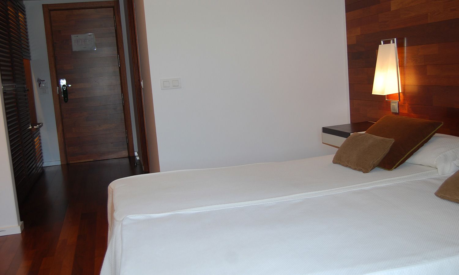Una habitación de hotel con una puerta de madera, una mesita de noche con lámpara y una cama con almohadas.