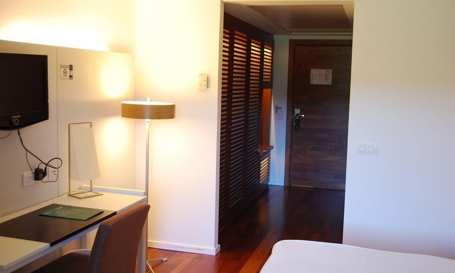A hotel room with a desk, chair, lamp, and a bed.