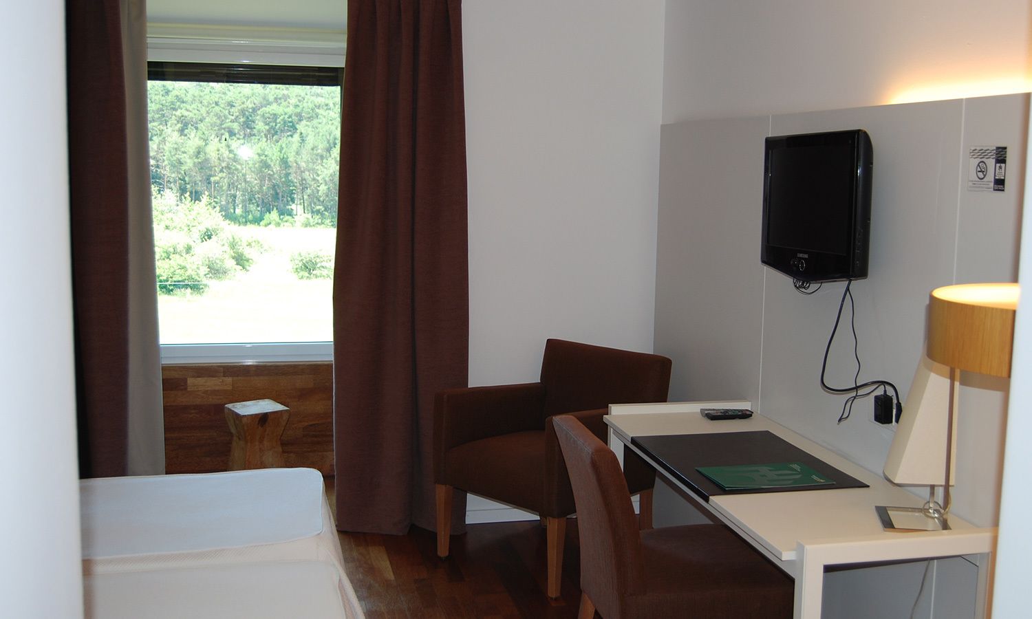 Una habitación de hotel con escritorio, silla, televisor, lámpara y ventana con cortinas.