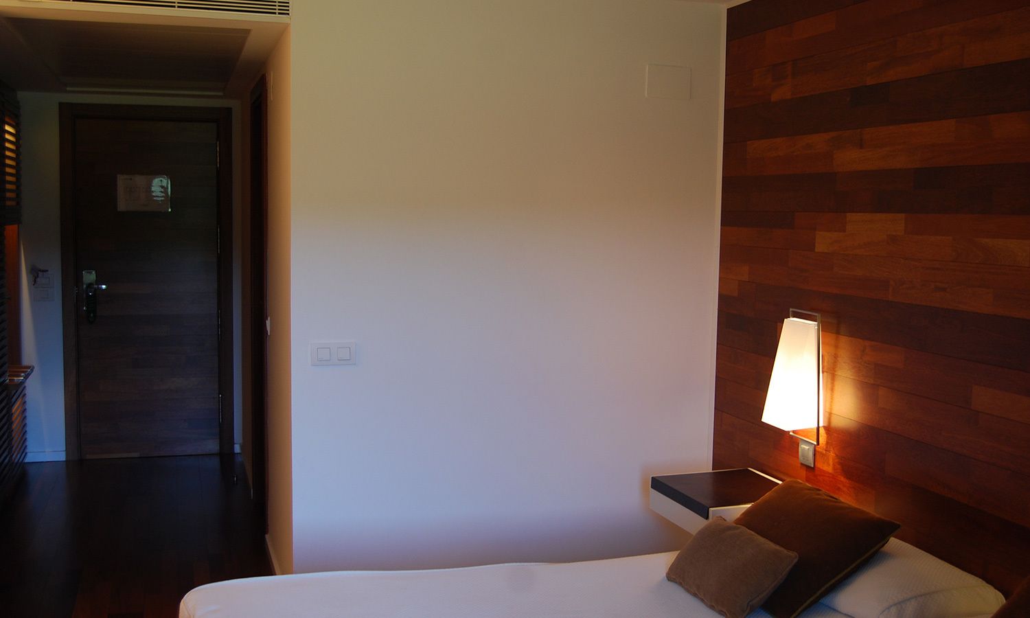 Habitación de hotel con cama, mesita de noche, lámpara de pared y panel de madera.