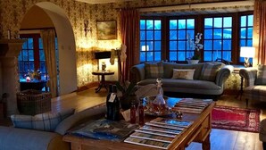 A cozy living room with a sofa, a coffee table with magazines, a fireplace, and a view of the outdoors through large windows. - Highland Bear Lodge & Woodland Bear Huts (Inverness)