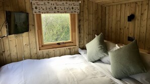 A wooden cabin bedroom with a bed, pillows, a window, and a wall-mounted TV. - Highland Bear Lodge & Woodland Bear Huts (Inverness)