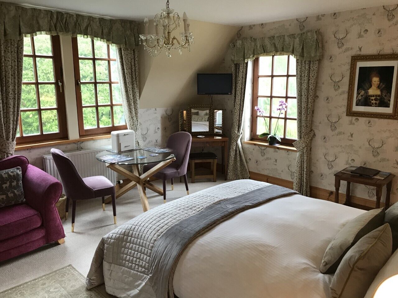 A bedroom with a large bed, a desk with a chair, a purple sofa, and a television on the wall.