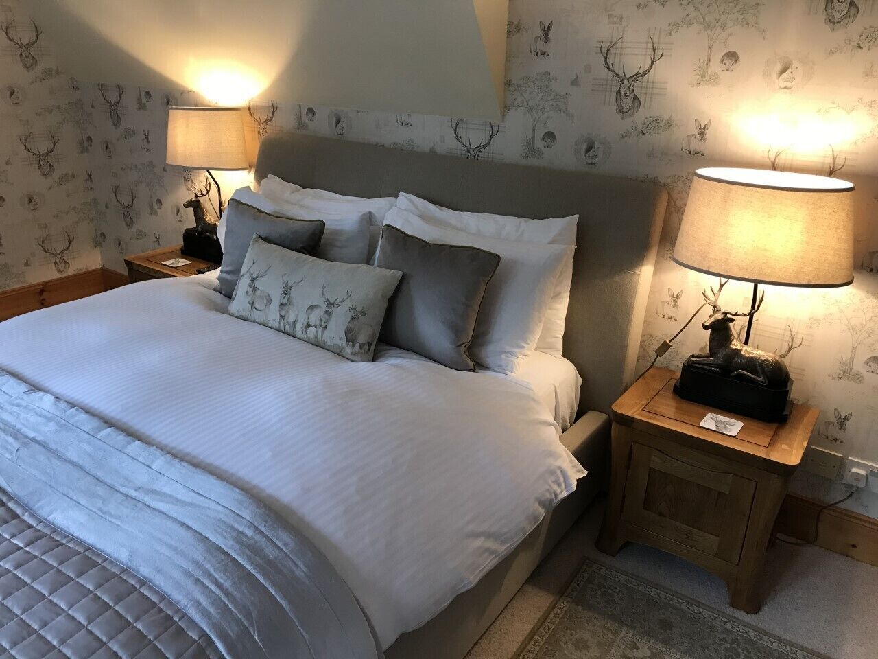 Un lit bien fait, avec une couette blanche et des oreillers gris, deux lampes de chevet et une petite table de nuit en bois sur laquelle se trouvent une lampe et un objet décoratif.