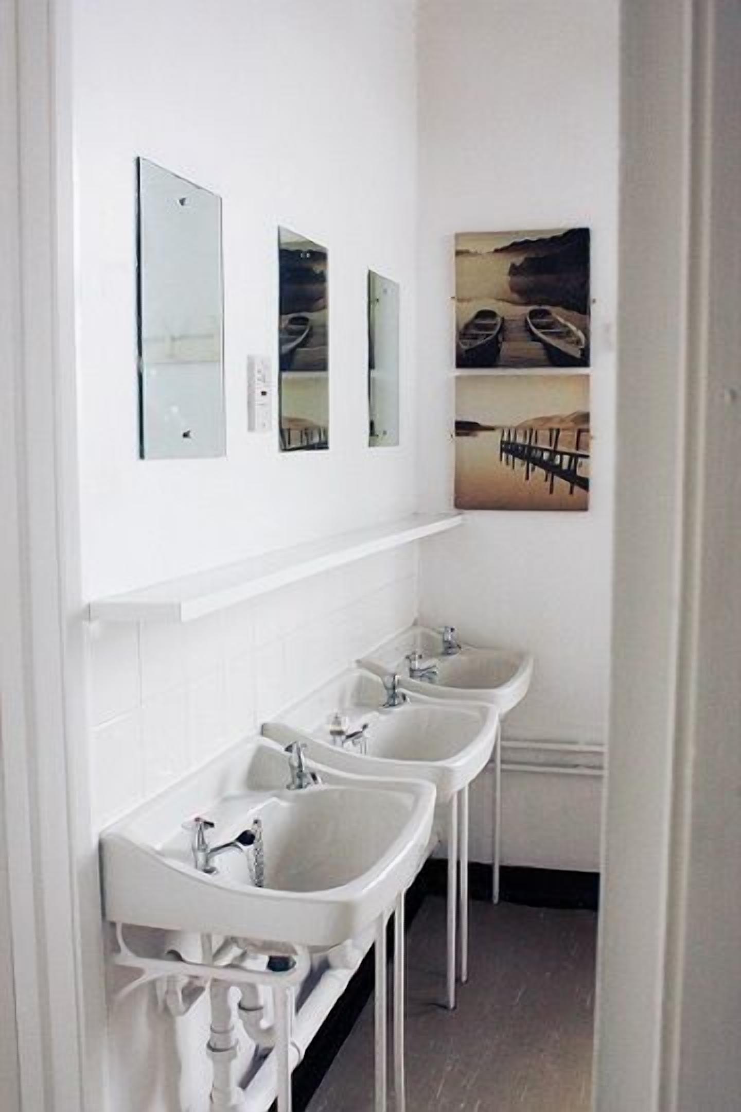 Un baño con tres lavamanos, paredes cubiertas de azulejos blancos y un cuadro de barcos en un muelle.