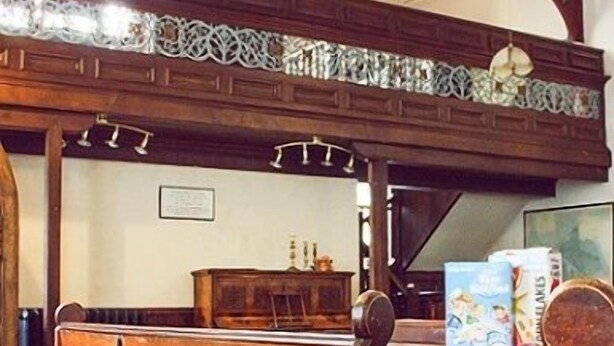 Un banco de madera con un tapete rojo estampado, un piano y una mesa con libros.