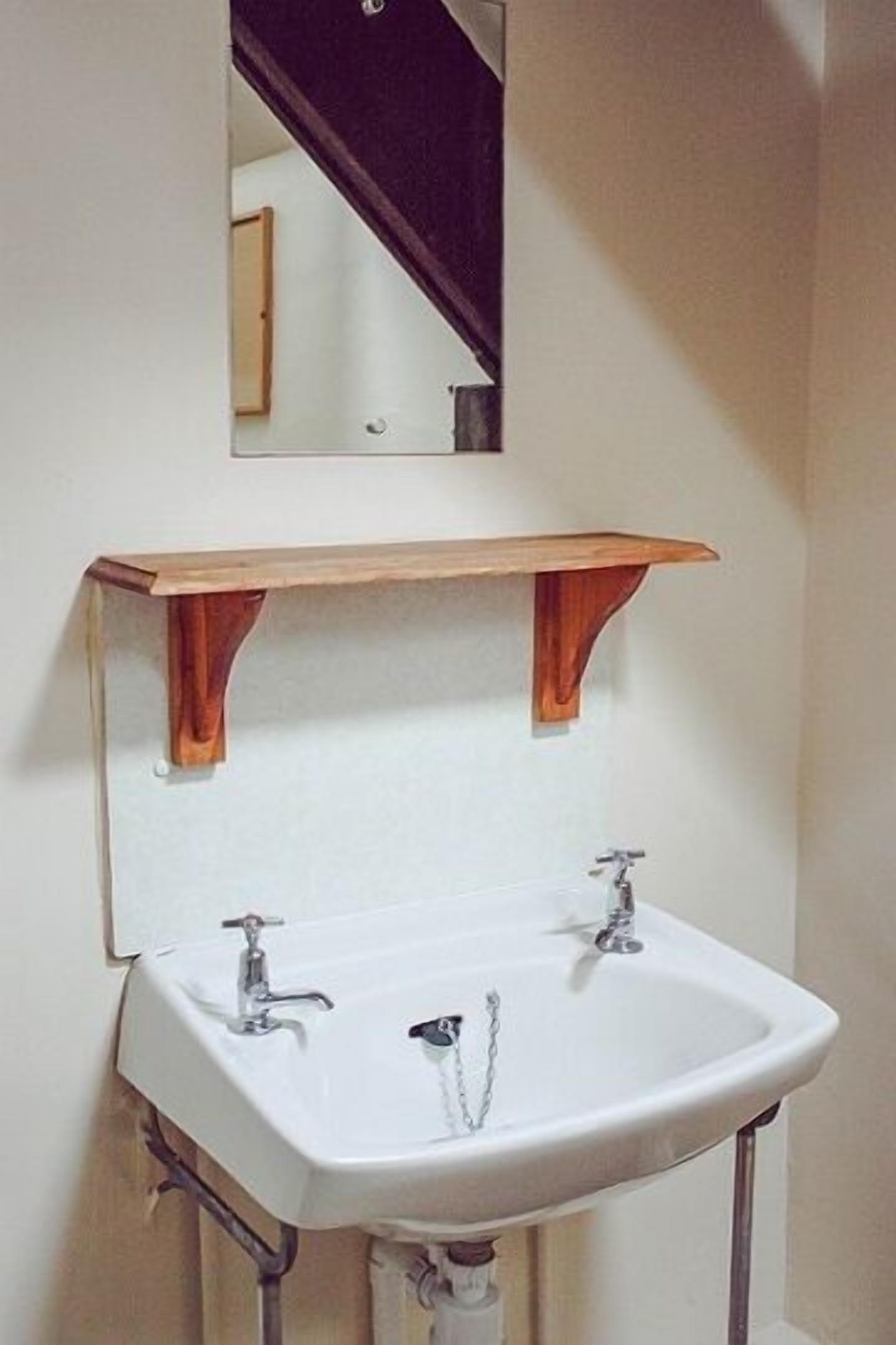 A bathroom with a white sink, two faucets, and a wooden shelf above it.