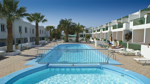 Une piscine à l’eau d’un bleu cristallin, entourée de palmiers et de bâtiments blancs dotés de balcons.