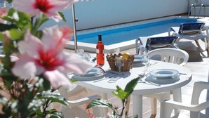 Mesa redonda blanca dispuesta para comer, con platos, cubiertos y una cesta de pan y limones. Se ve una piscina al fondo.