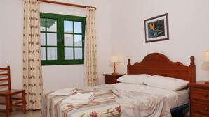 Un dormitorio con cama, una silla, una mesita de noche, una lámpara y un ventanal con cortinas.