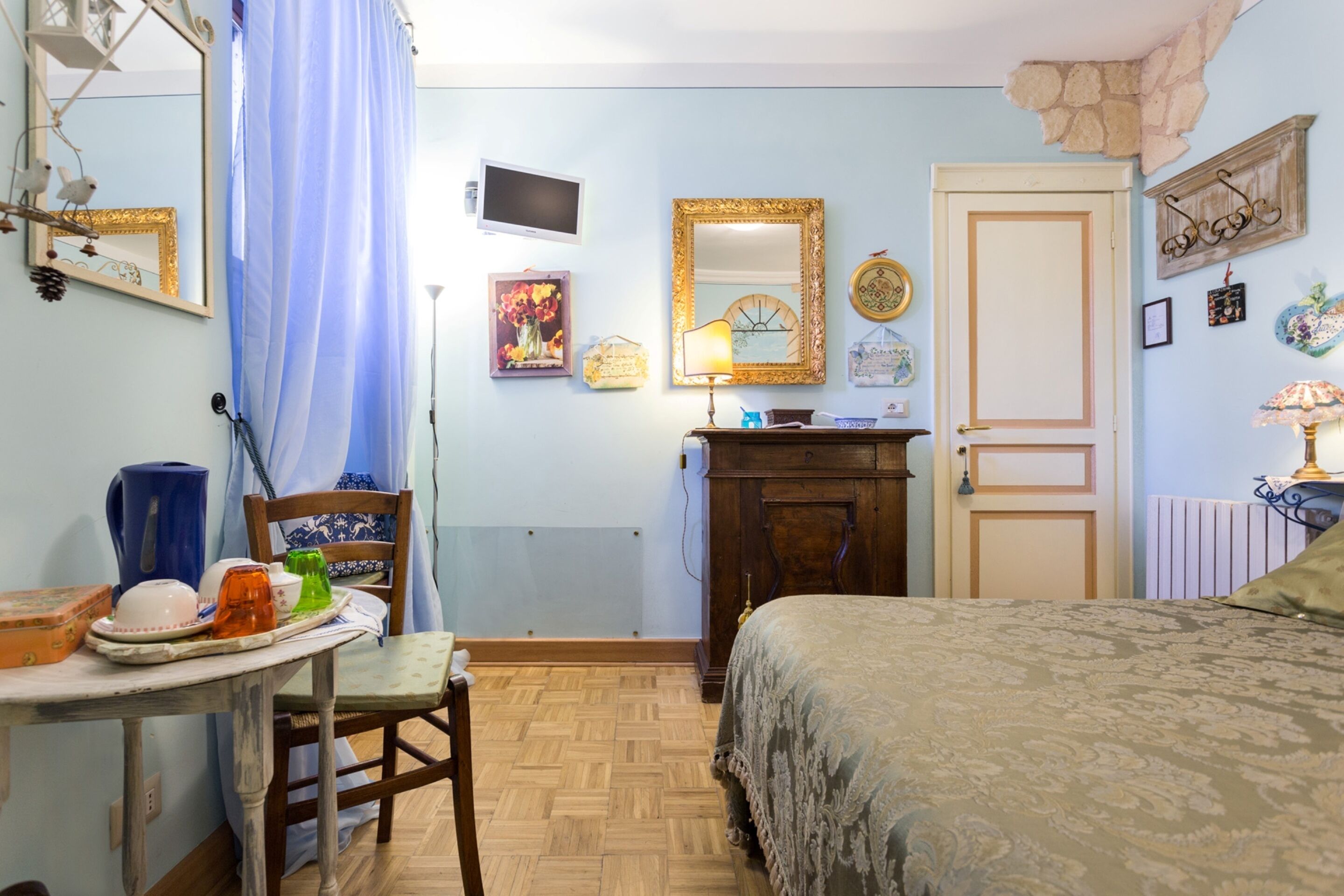 A bedroom with a bed, a dresser, a chair, a table with items, a mirror, a television, and a door.