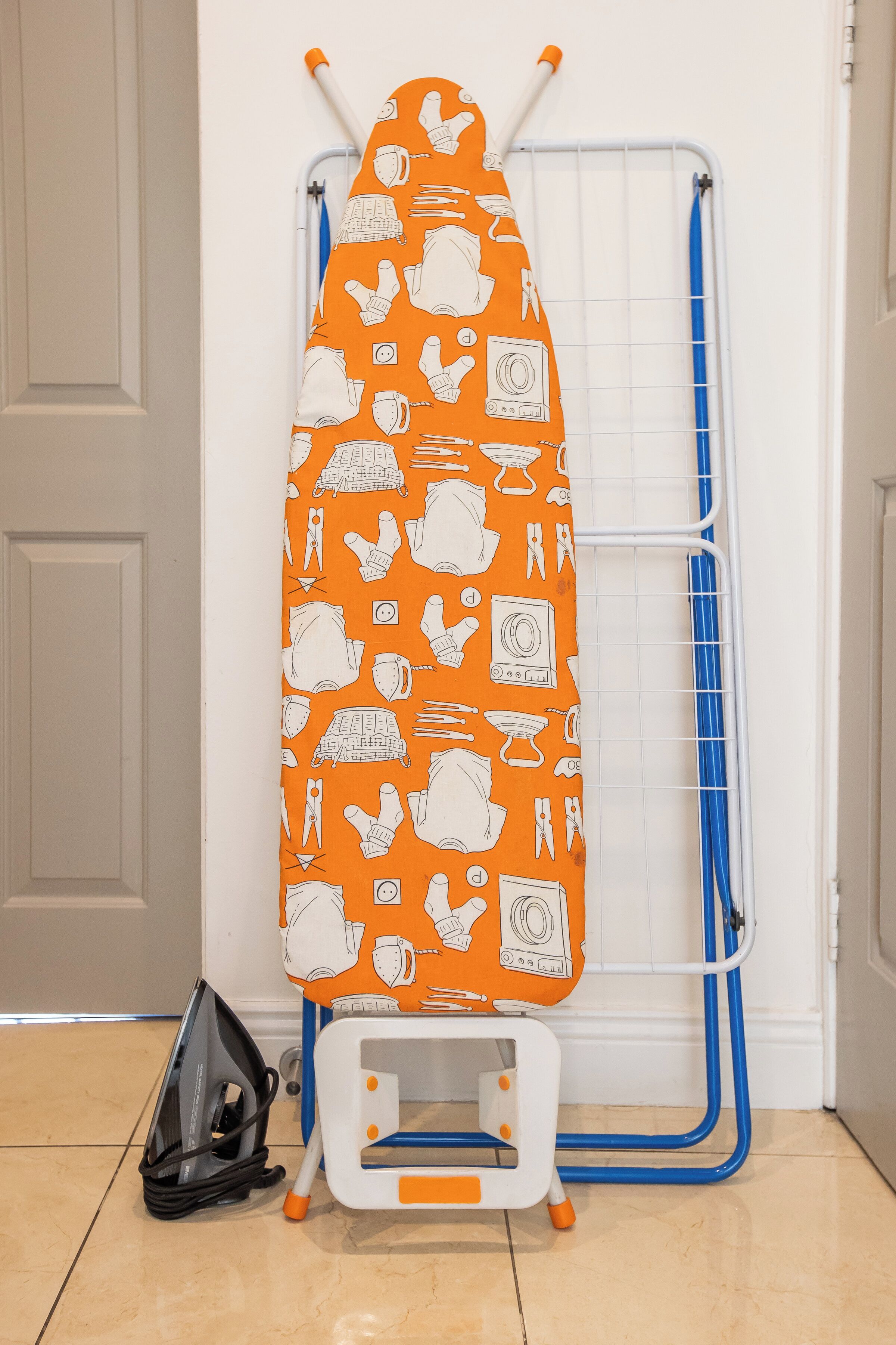 A portable ironing board with an orange and white pattern, a small ironing board stand, and a black bag on a tiled floor.