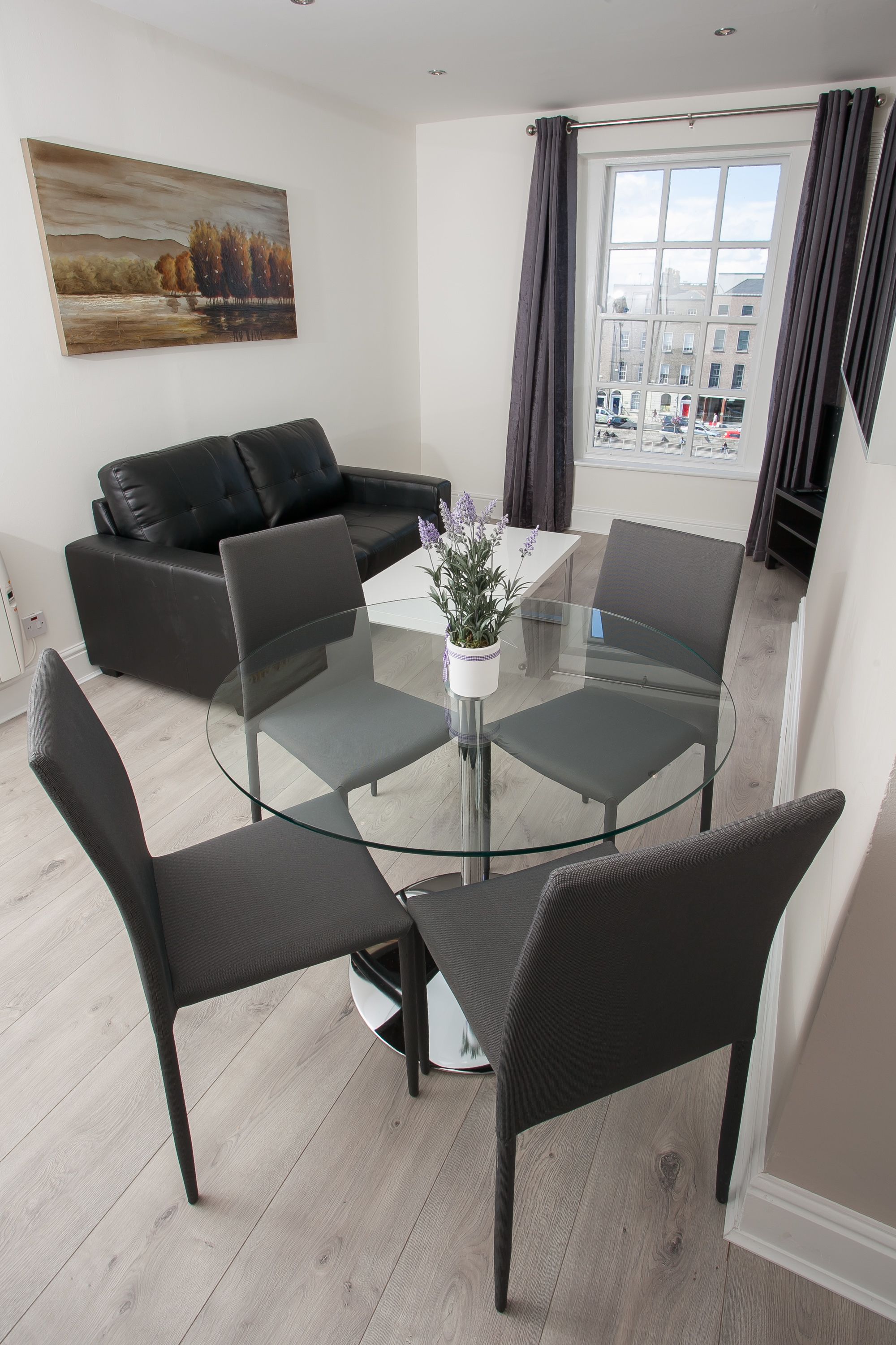 A modern living room with a glass table surrounded by chairs, a black leather sofa, and a painting on the wall.