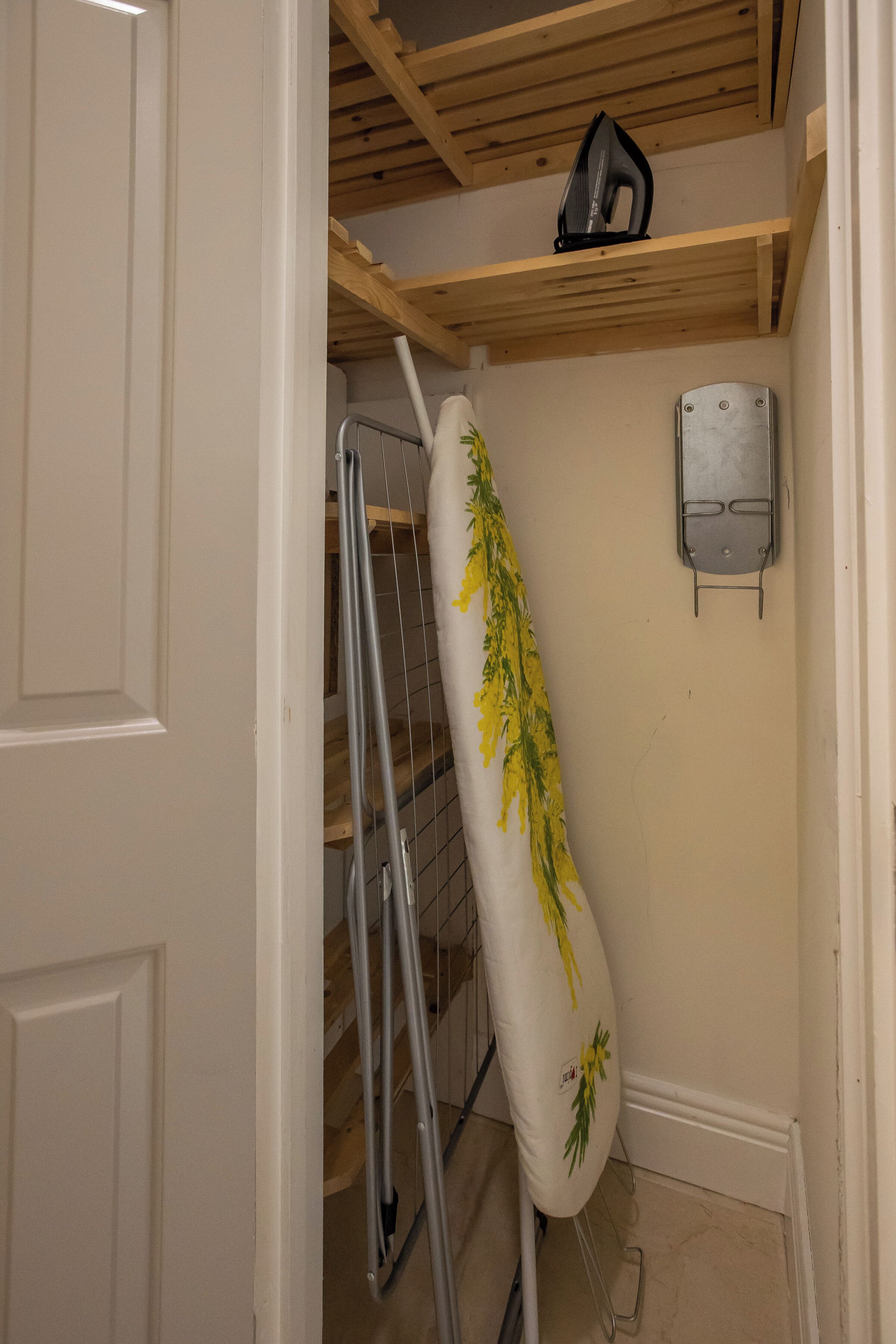 Une pièce avec une porte blanche, un plafond en bois, une planche à repasser blanche ornée de motifs floraux, un support en métal pour la planche à repasser et une prise électrique fixée au mur.