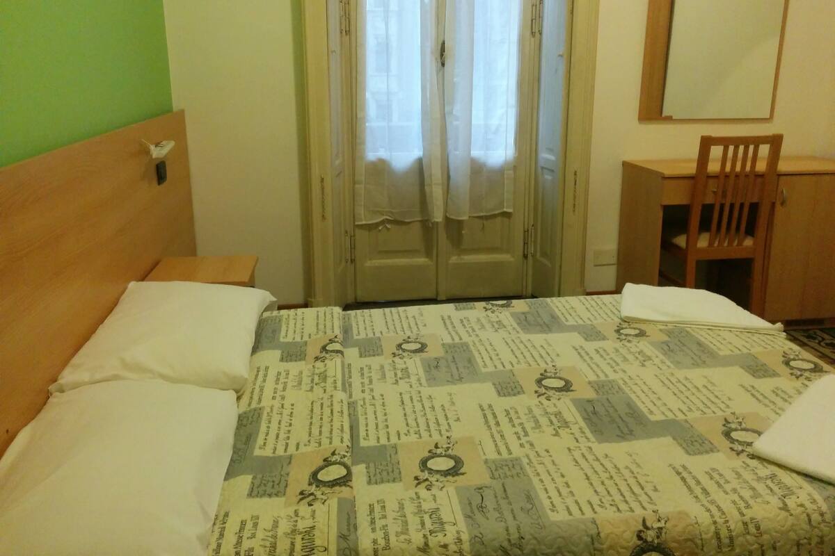 Um quarto com cama, cabeceira de madeira, escrivaninha com cadeira, espelho e guarda-roupa.