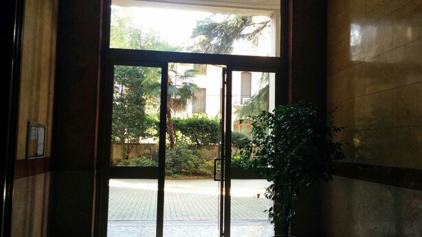 Une entrée avec une porte en verre donnant sur un espace pavé et une zone verdoyante à l’extérieur.