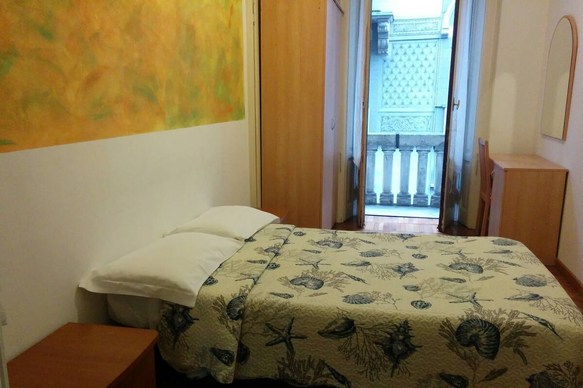 Quarto com cama, mesa de cabeceira de madeira e porta que dá para uma varanda.