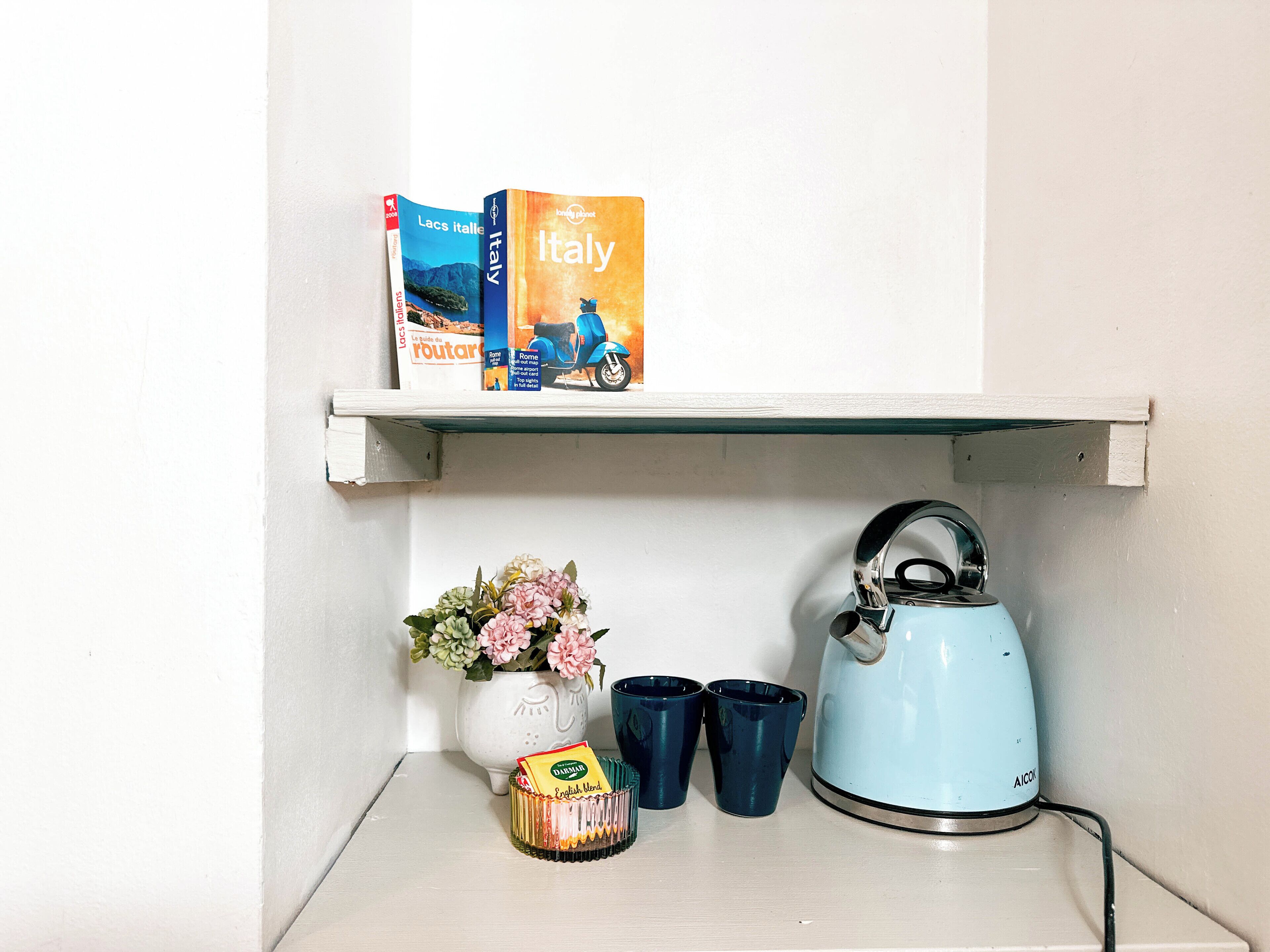 Una estantería con libros de viajes, un florero con flores, una tetera y dos tazas.