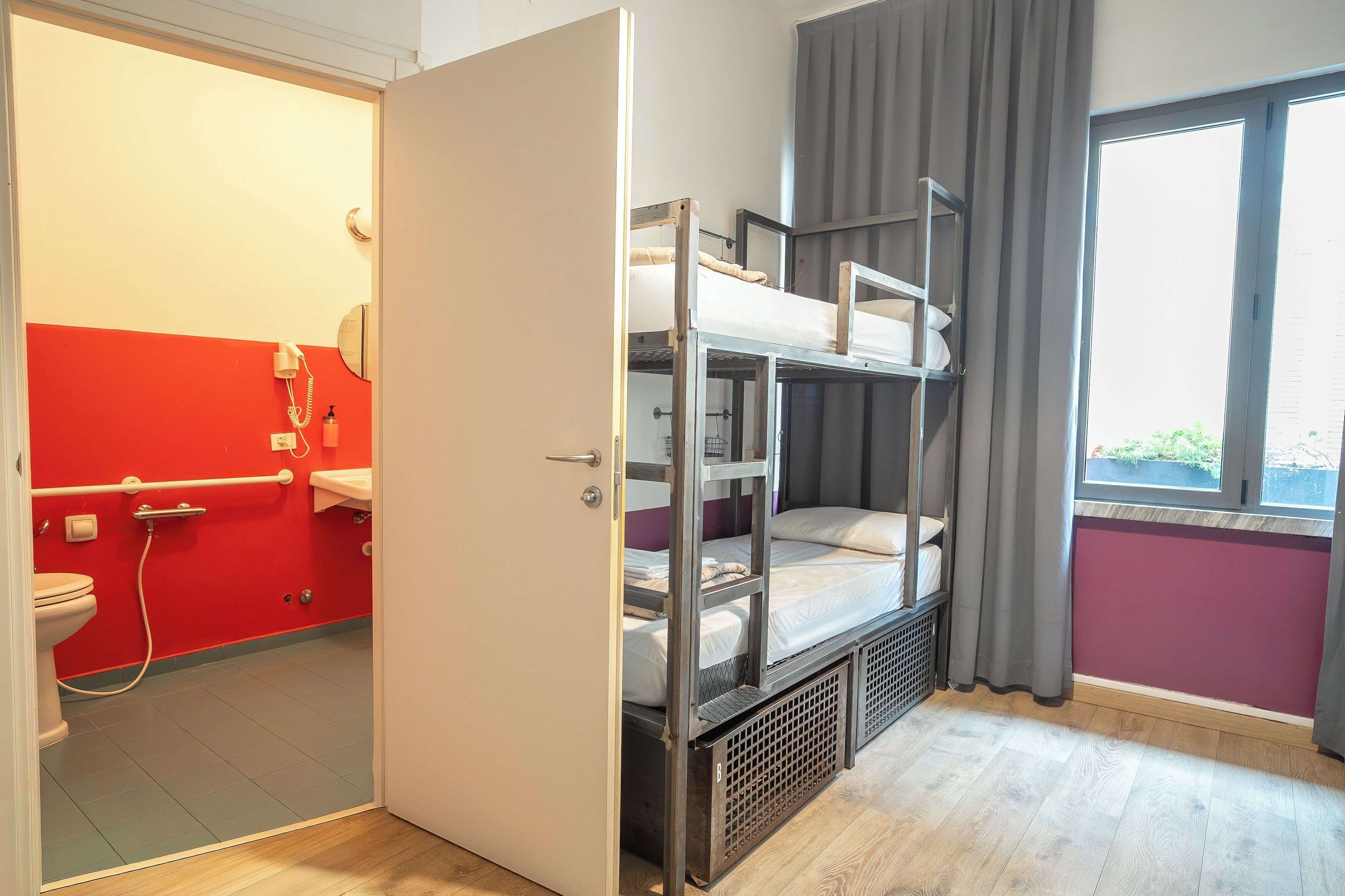 A room with bunk beds, a bathroom with a red wall, and a window with curtains.