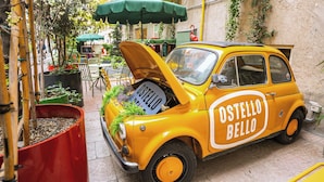 A yellow vintage car with 'Ostello Bello' written on its side parked in a narrow street with outdoor seating and greenery. - Ostello Bello Milano Duomo (Milan)