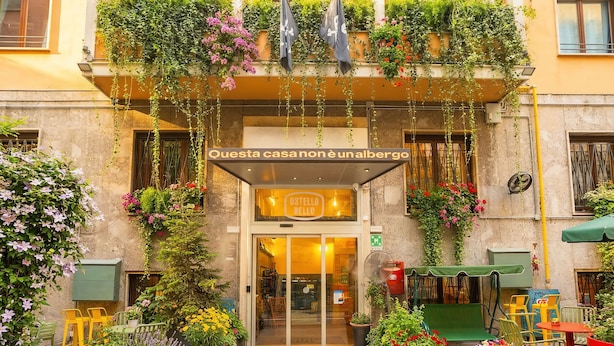 A hotel entrance with a sign 'Ostello Bello' and a glass door. There are outdoor seating areas with chairs and tables, surrounded by potted plants and flowers. - Ostello Bello Milano Duomo (Milan)