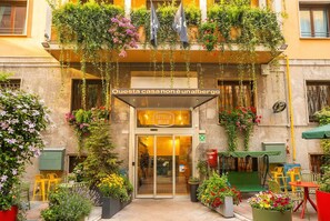 A hotel entrance with a sign 'Ostello Bello' and a glass door. There are outdoor seating areas with chairs and tables, surrounded by potted plants and flowers. - Ostello Bello Milano Duomo (Milan)
