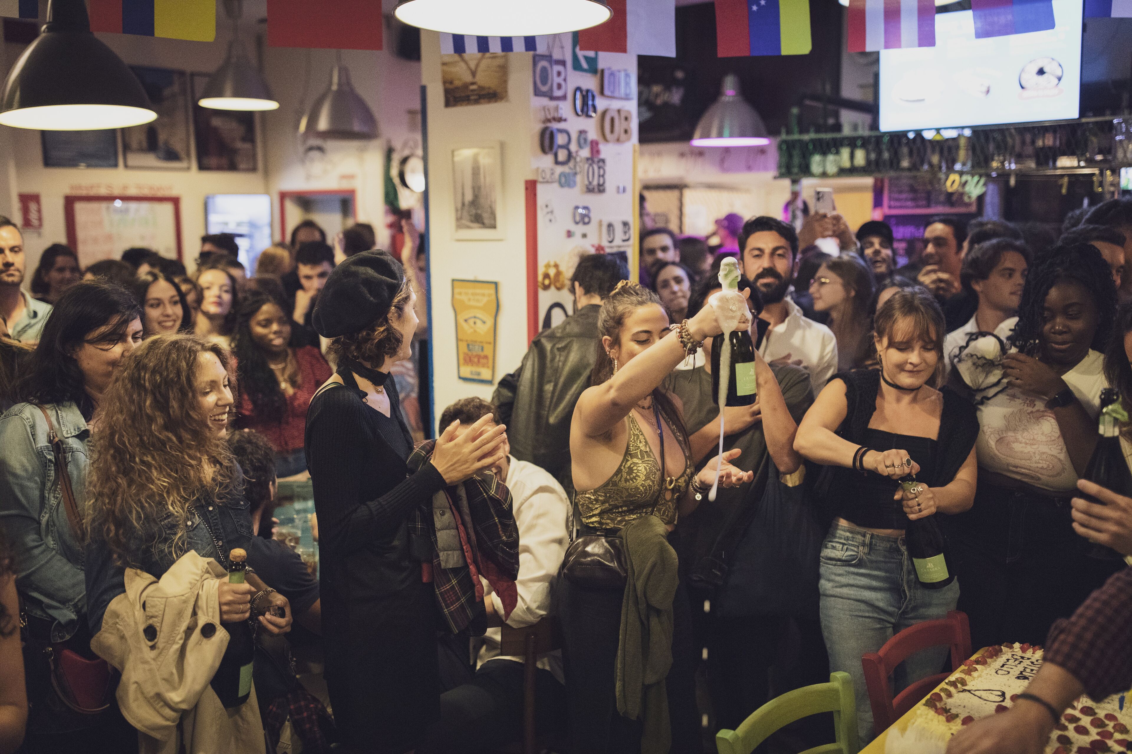 A crowded indoor space with people gathered around tables, some holding drinks.