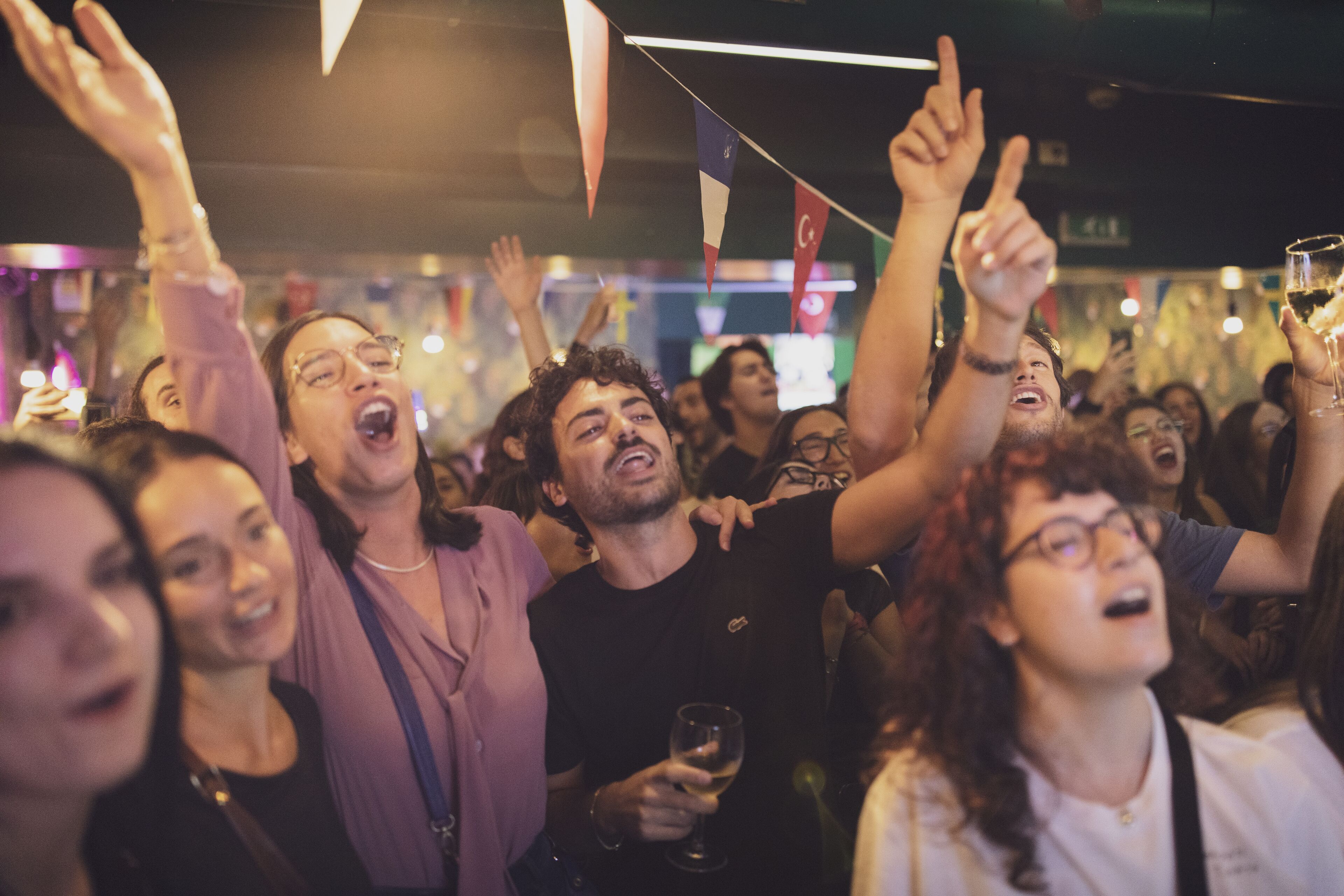 A crowded indoor event with people raising their hands and holding drinks.