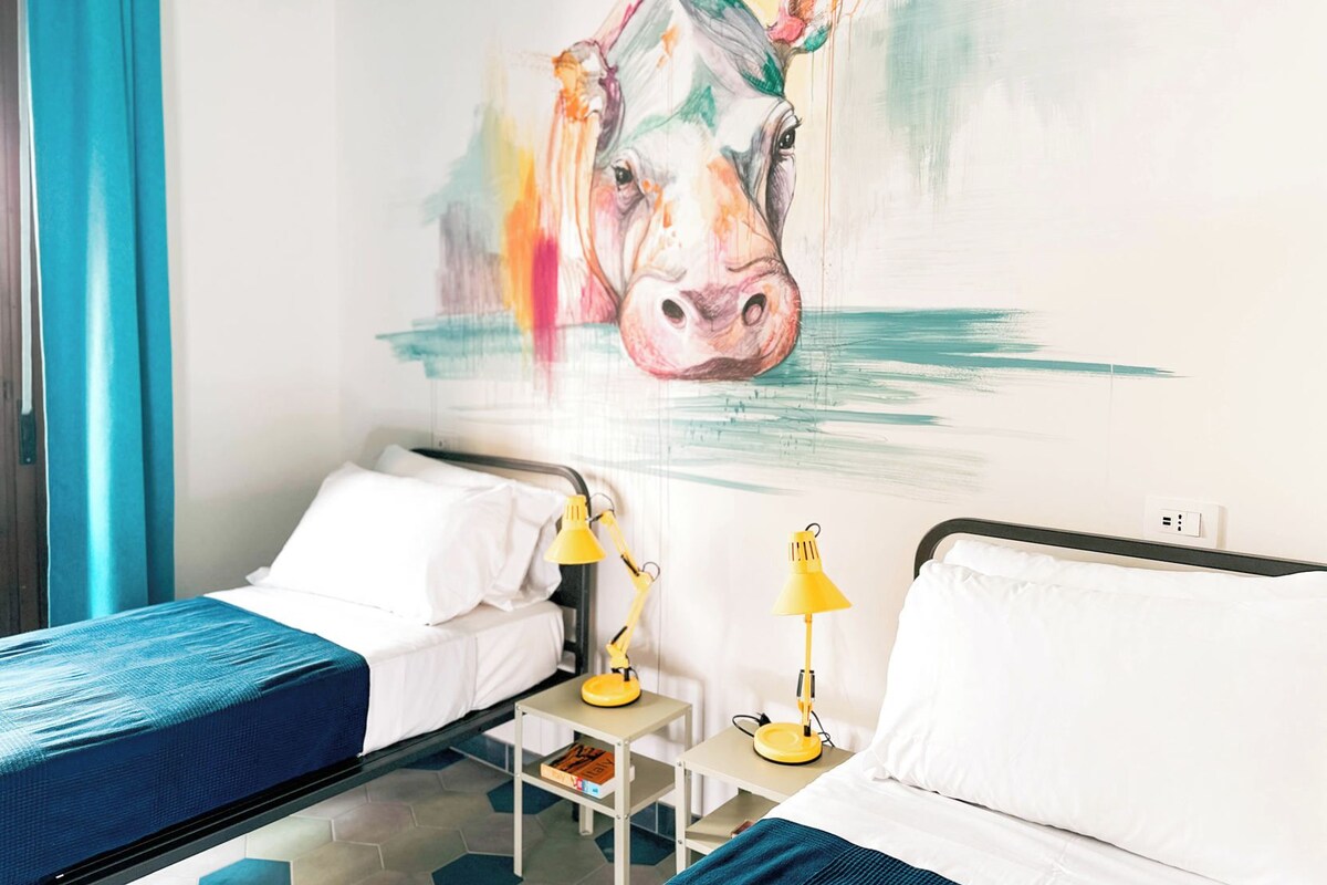 A hotel room with two beds, a painting of a cow on the wall, and a small table with a lamp.