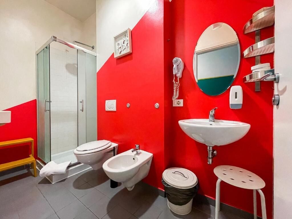 A bathroom with a red wall, a glass shower enclosure, a toilet, a bidet, a sink, a mirror, and a trash bin.