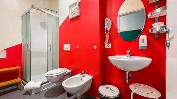 Ein Badezimmer mit roter Wand, einer Duschkabine aus Glas, einer Toilette, einem Bidet, einem Waschbecken, einem Spiegel und einem Mülleimer.