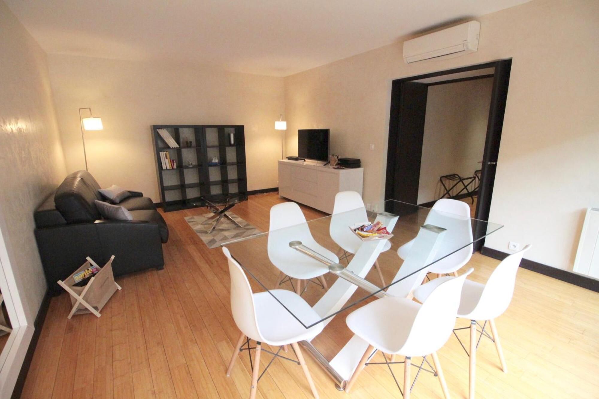 A modern living room with a glass dining table, white chairs, a black sofa, and a bookshelf.