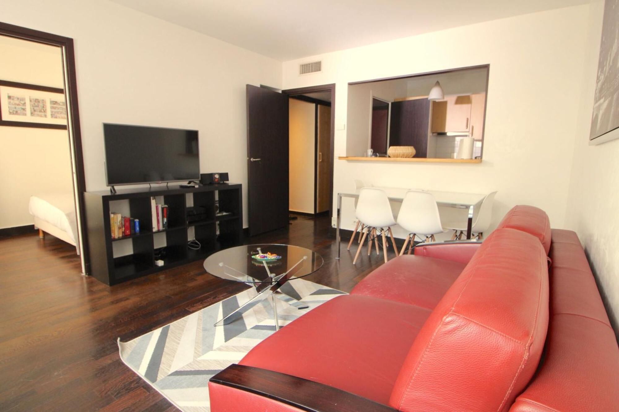 Een moderne woonkamer met een rode leren bank, een glazen salontafel en een zwarte tv-kast met een flatscreen televisie. Er is een eethoek met een witte tafel en stoelen.