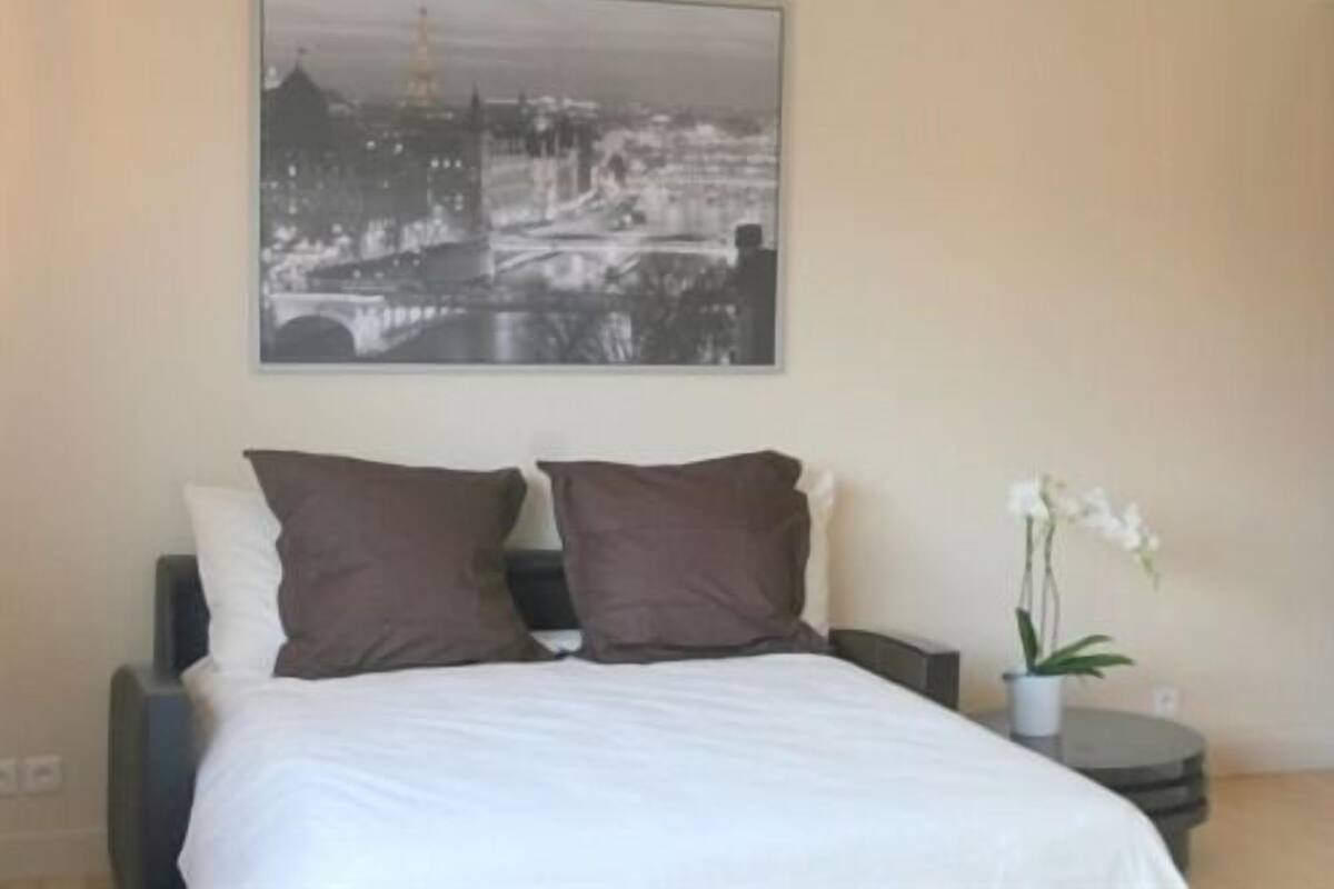 Un dormitorio con cama, dos almohadas, una mesita de noche, una orquídea en maceta y una pintura de paisaje urbano en la pared.