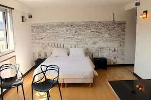 Een slaapkamer met een bed, twee stoelen, een nachtkastje en een muurschildering van een stadsgezicht.