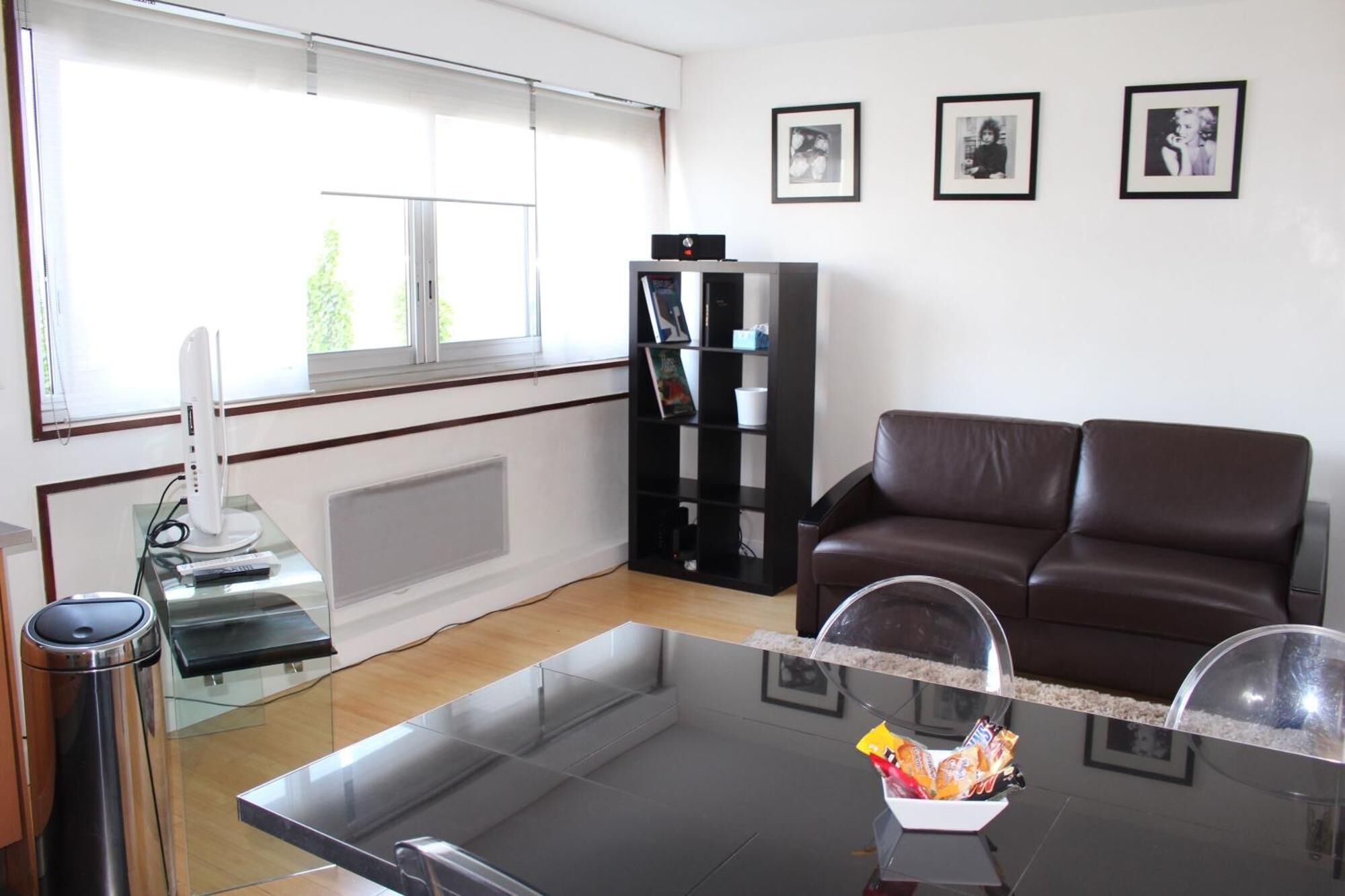 A living room with a glass coffee table, a brown leather sofa, a bookshelf, and a window with blinds.