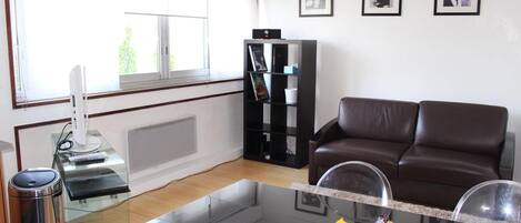 A living room with a glass coffee table, a brown leather sofa, a bookshelf, and a window with blinds.