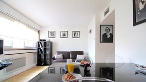 En moderne stue med et sofabord i glas, en sofa og en bogreol. Vægtene er dekoreret med billeder, og et vindue har skodder.