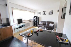 Ein modernes Wohnzimmer mit einem Glastisch, einer Couch und einem Fernseher. An den Wänden hängen Bilder in Rahmen, und es gibt eine Küchenzeile mit Mikrowelle und Toaster.