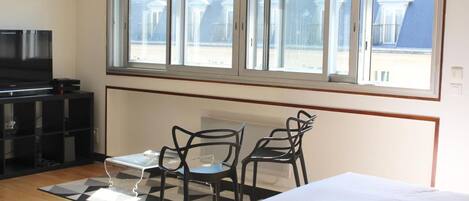 A hotel room with a bed, a TV, a small table with chairs, and a window with a view of buildings.