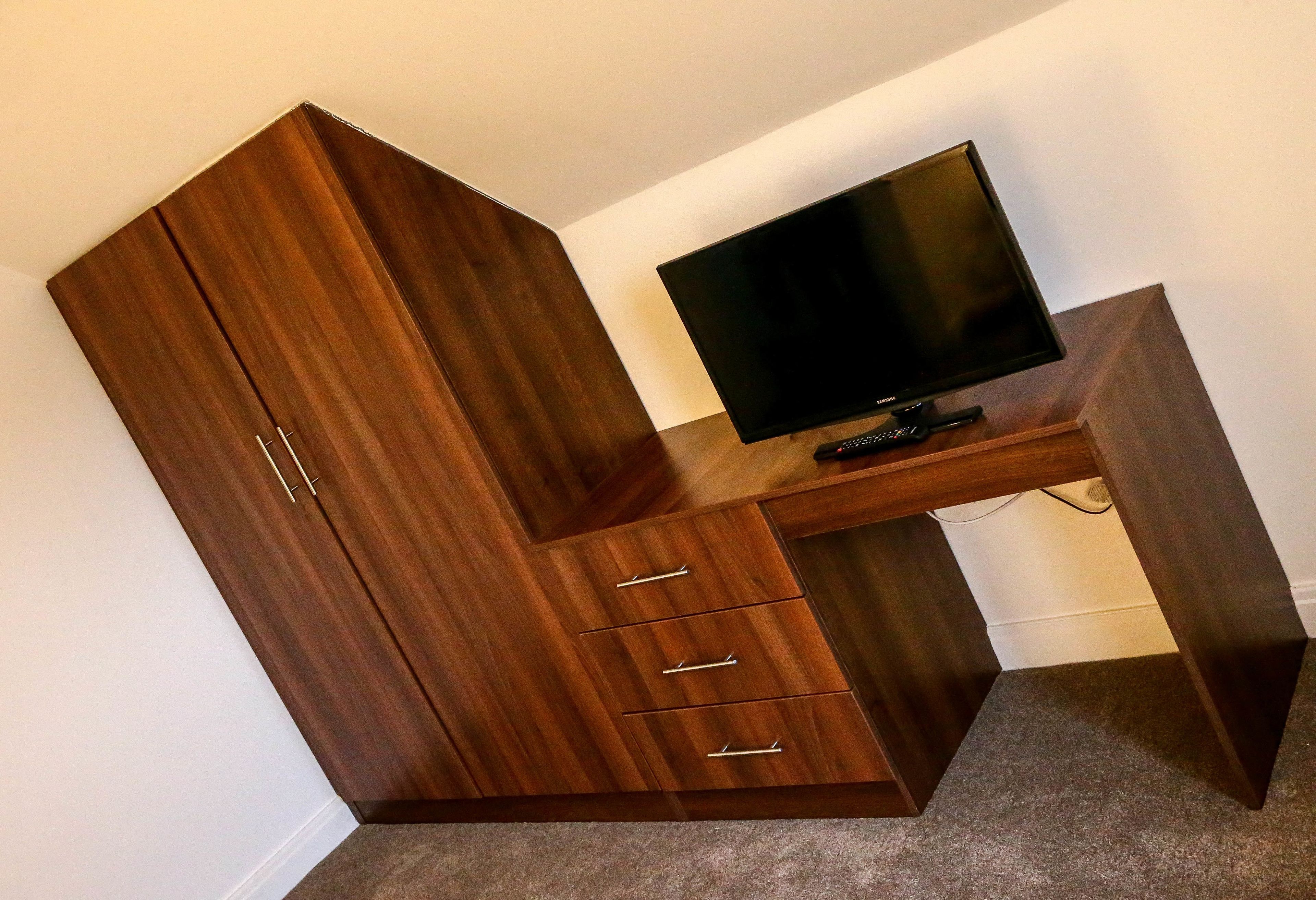 Habitación de esquina con escritorio de madera y televisor, que incluye un armario con tres cajones y dos puertas.