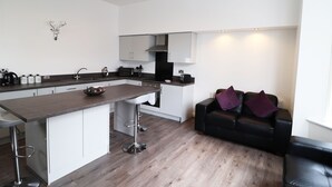 A modern kitchen with a central island, bar stools, and a black leather sofa in a living area. - Breck Blackpool (Blackpool)