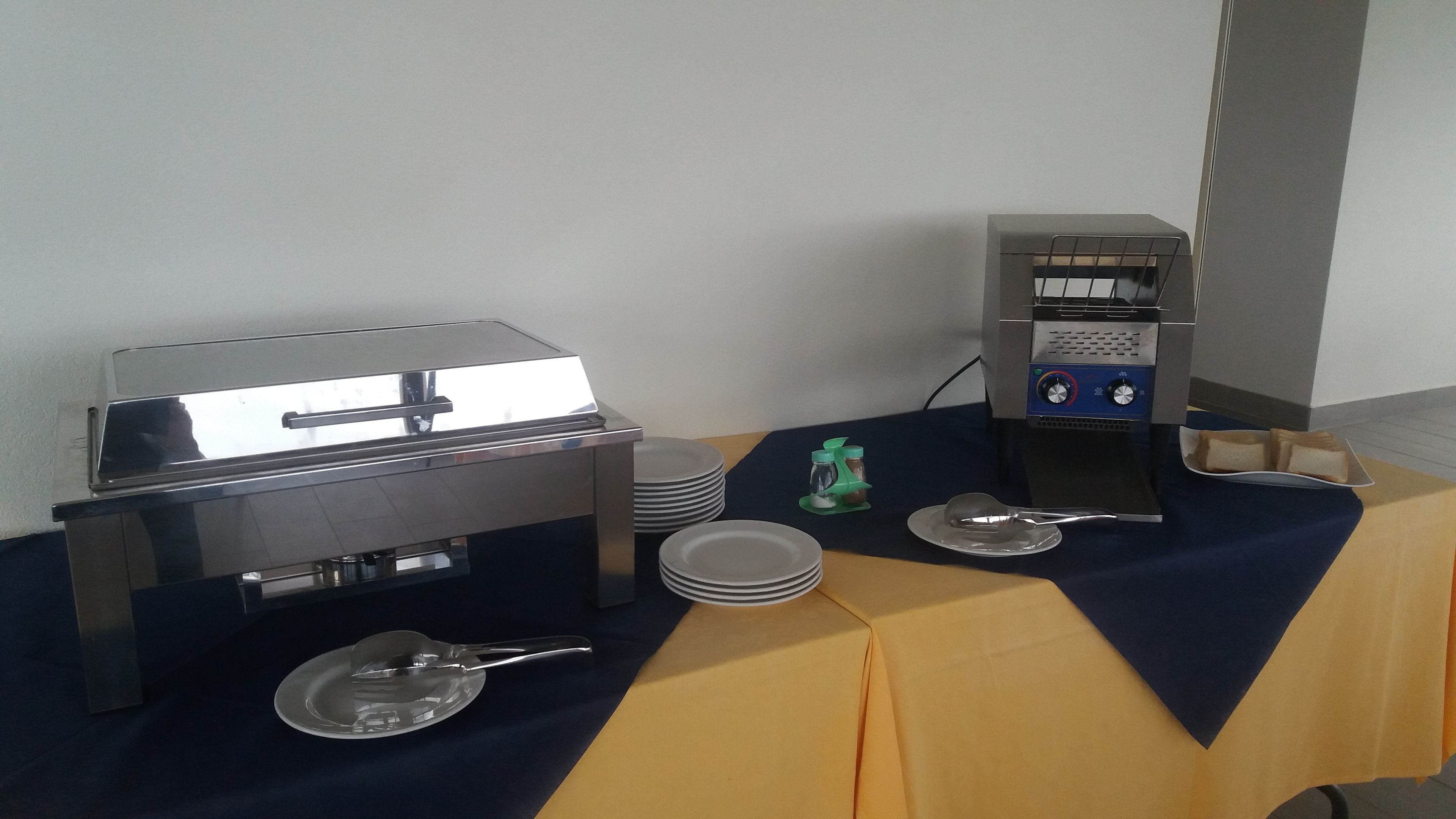 A buffet setup with a grill, a coffee machine, and stacked plates on a table covered with a blue tablecloth.