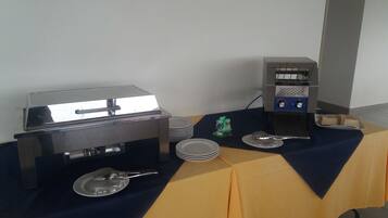 A buffet setup with a grill, a coffee machine, and stacked plates on a table covered with a blue tablecloth.