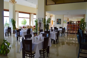 Uma sala de jantar ampla com várias mesas e cadeiras, janelas grandes e plantas dentro de casa.